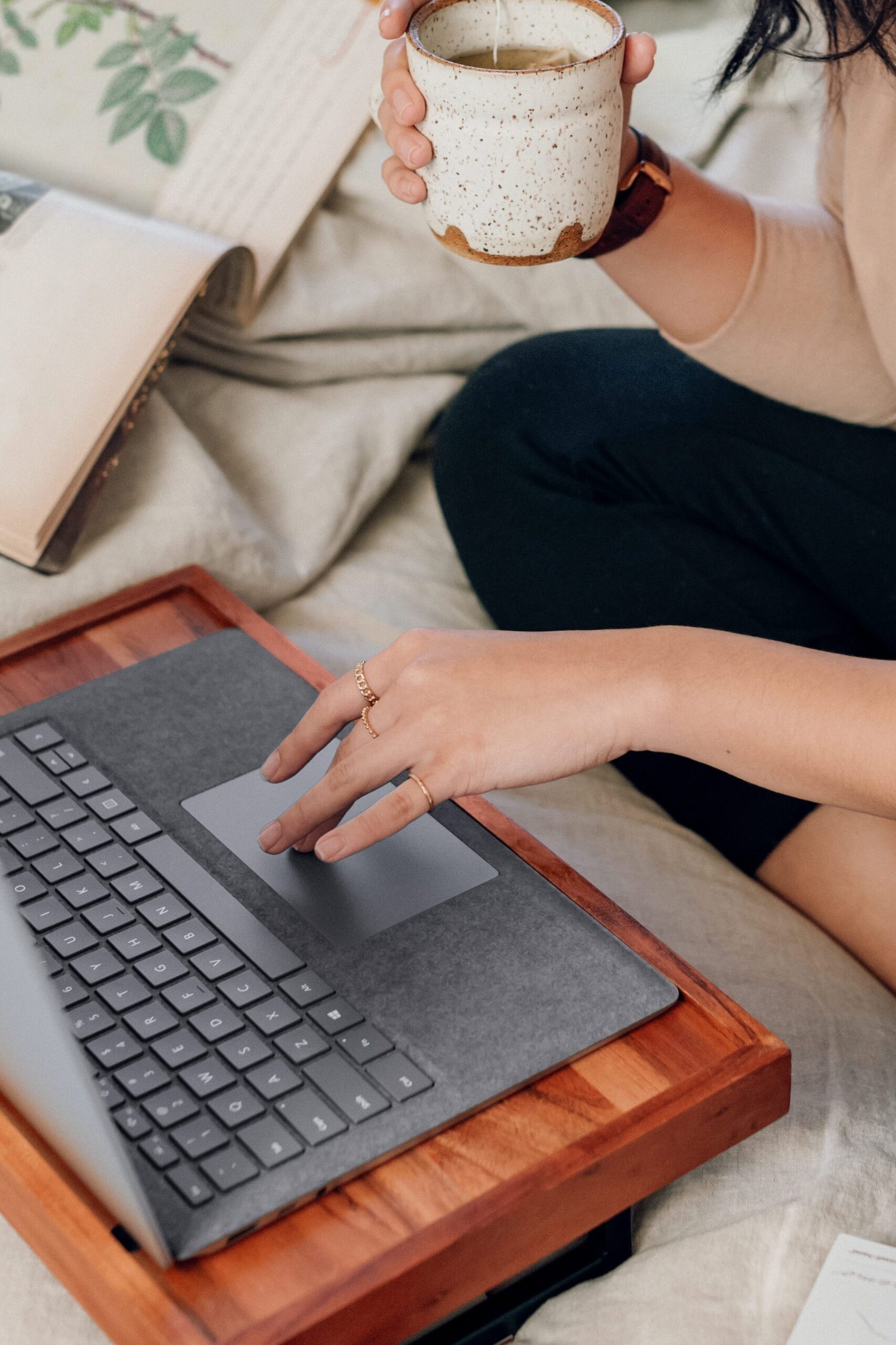 Mulher sentada na cama com seu computador. Ela digita com uma mão, e, com a outra, bebe um chá, como se estivesse trabalhando com as normas da ABNT.